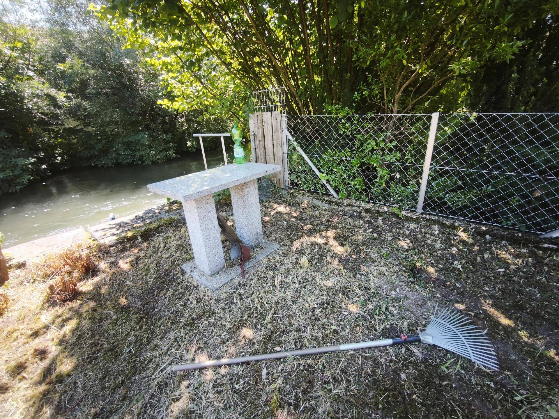 Steintisch im Gartenbereich mit einer Harke und üppiger Vegetation im sonnigen Hintergrund.