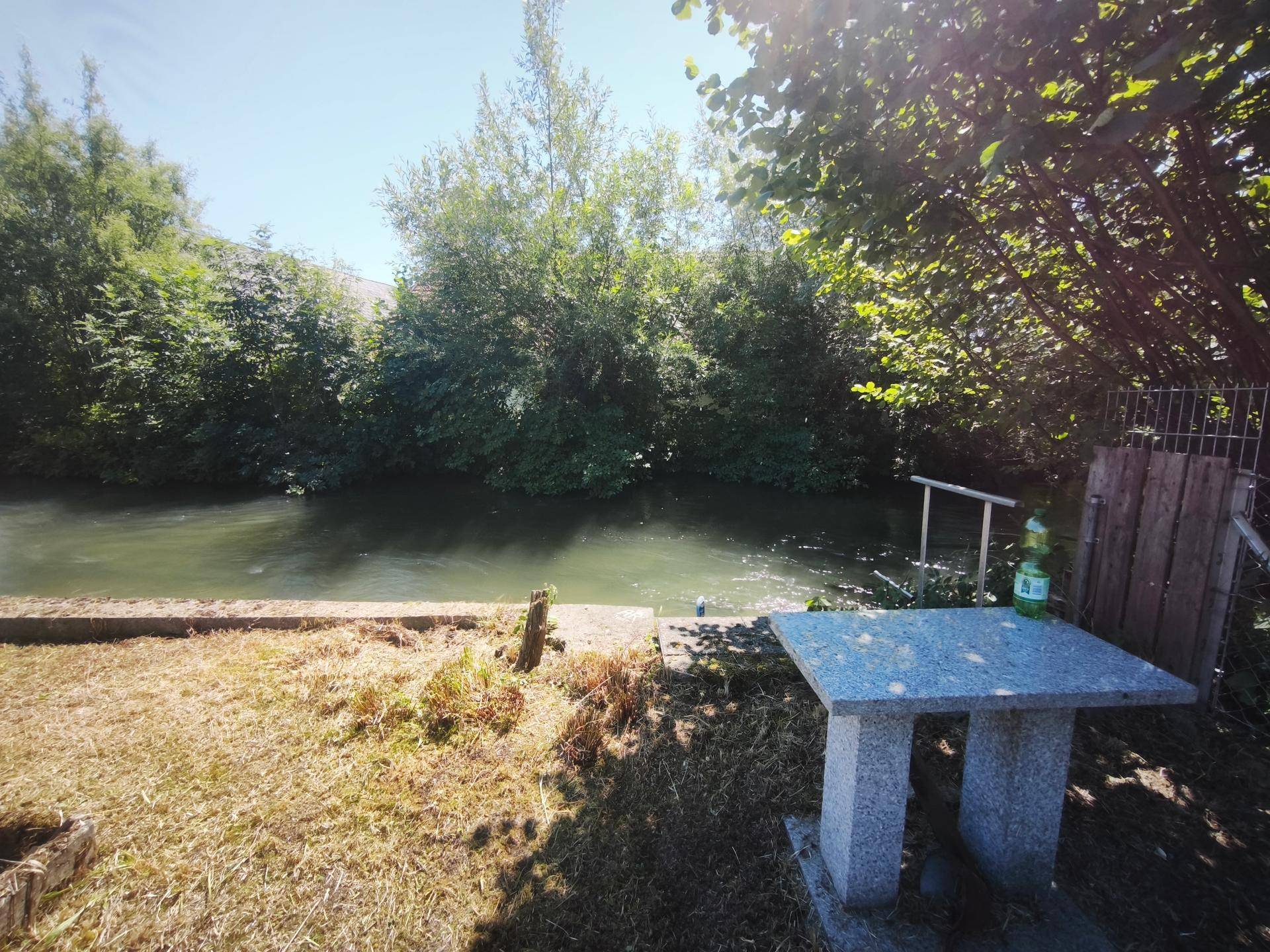 Steintisch am Flussufer mit Blick auf das Wasser und die umliegenden Bäume im Sonnenlicht.