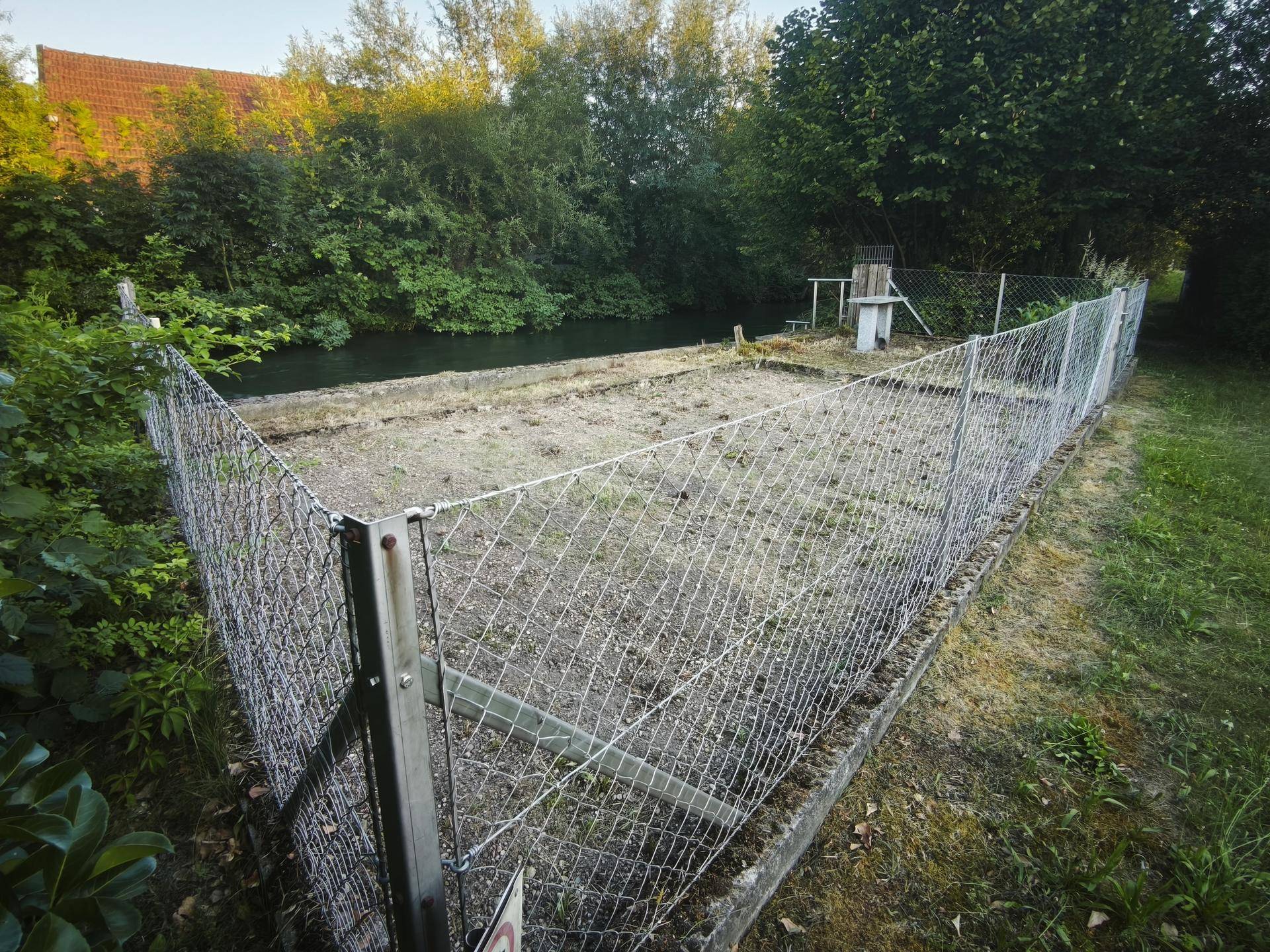 Eingezäunter Gartenbereich am Fluss mit einem kleinen Steintisch und üppiger Vegetation.