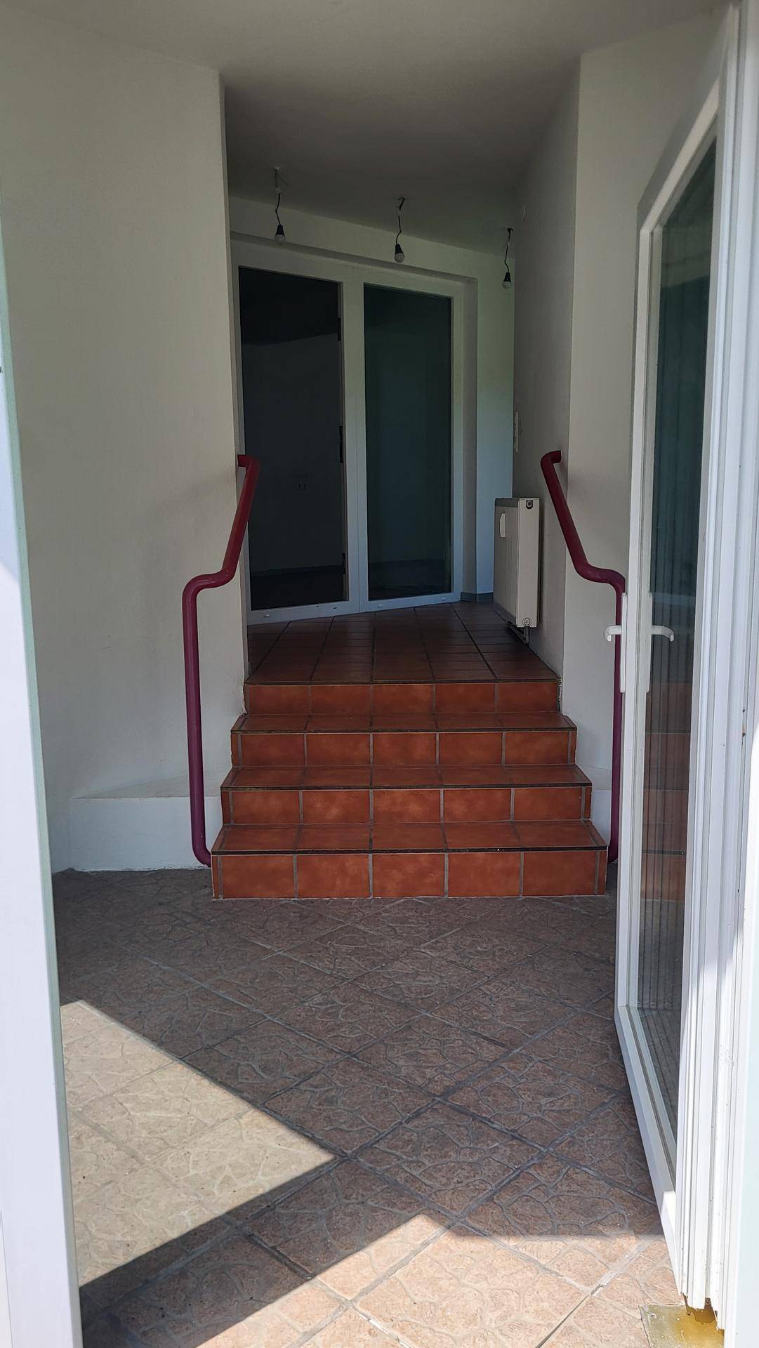 Eingangsbereich mit gefliestem Boden, Treppe und roten Handläufen, führt zu einer weiteren Tür.