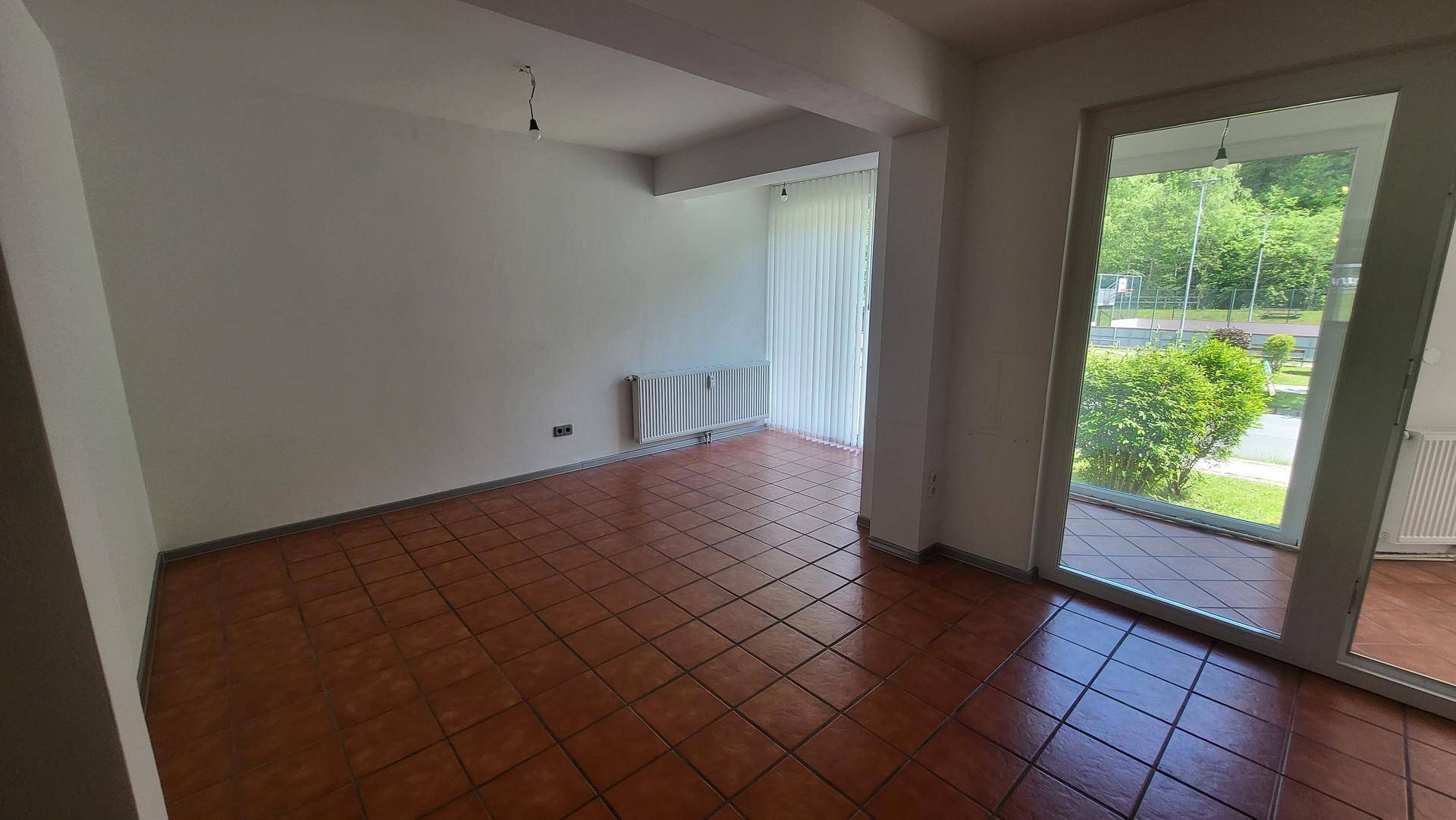 Heller Wohnraum mit großen Fenstern, braunen Fliesen und einem Heizkörper an der Wand.
