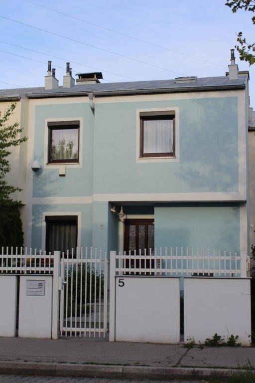 Reihenhausfassade in Hellblau mit weißen Akzenten, dunklen Fenstern und einem Vorgartenzaun.