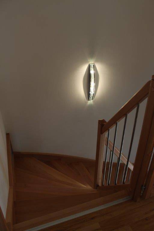 Holztreppe mit Metallgeländer und einer modernen Wandleuchte, die den Aufgang beleuchtet.