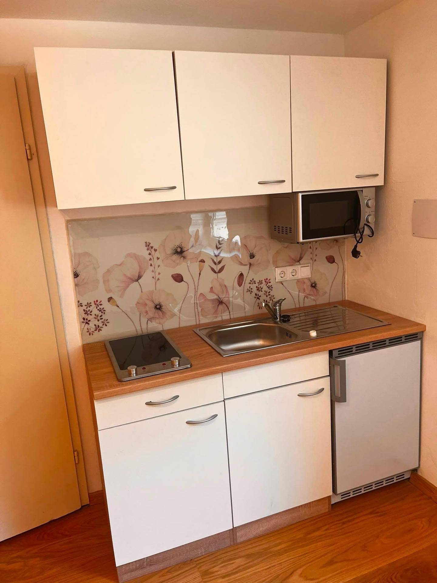 Kompakte Kochnische mit weißen Schränken, Induktionskochfeld und floralem Spritzschutz.