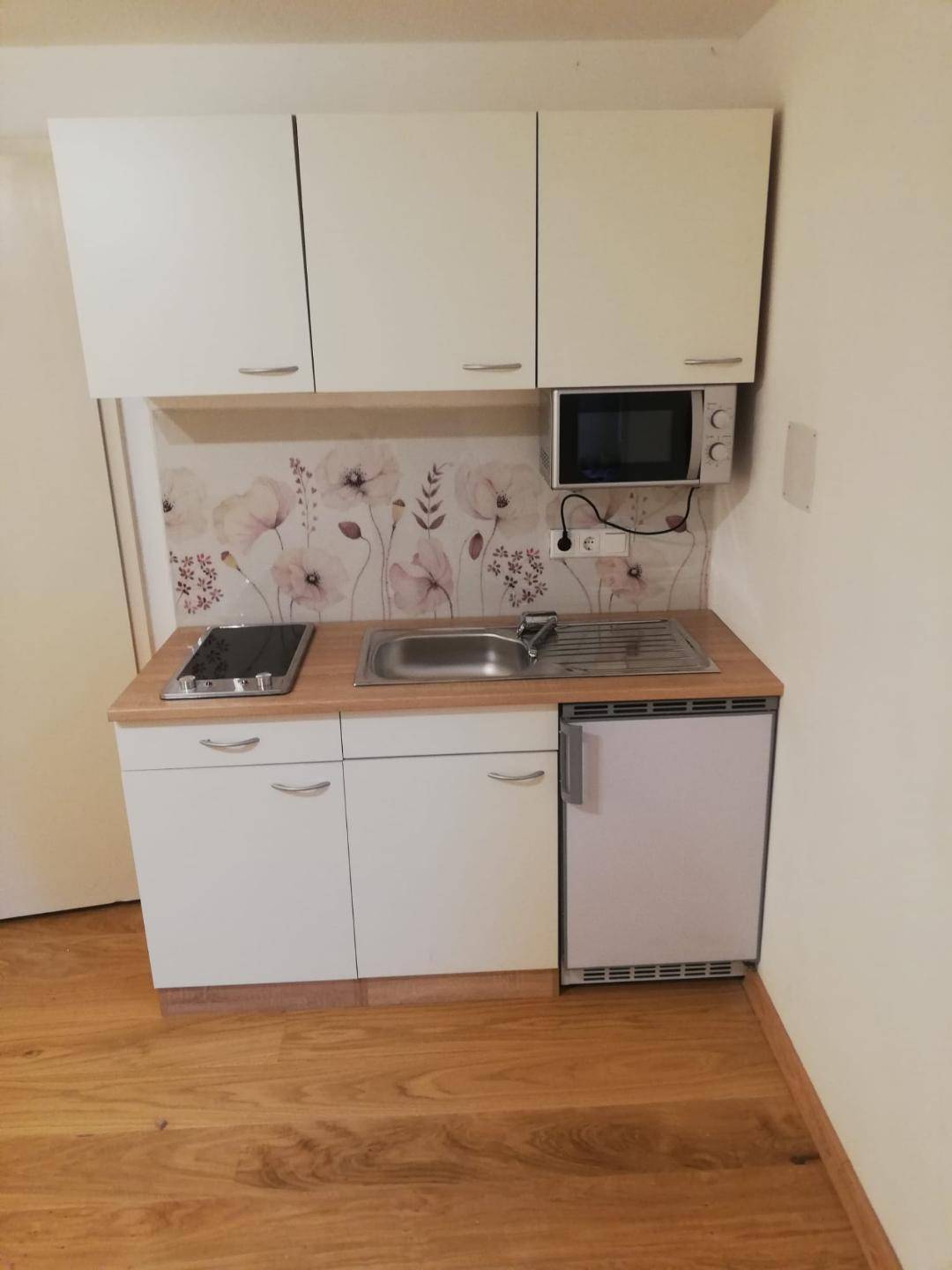 Kompakte Küchenzeile mit weißen Schränken, Spüle, Mikrowelle und Kühlschrank.