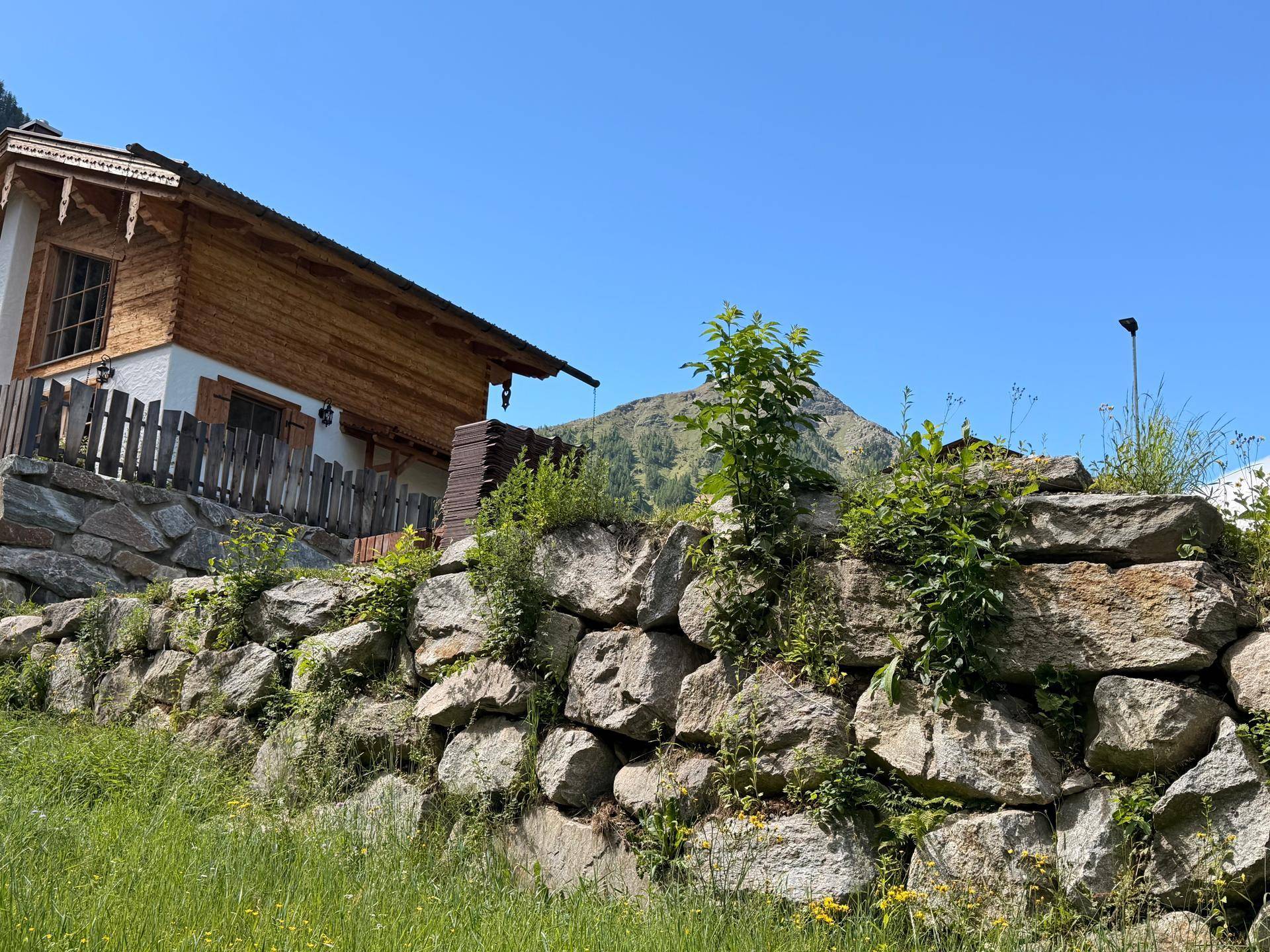 Traditionelles Holzchalet mit Steinmauer und weitem Bergblick an einem sonnigen Tag.