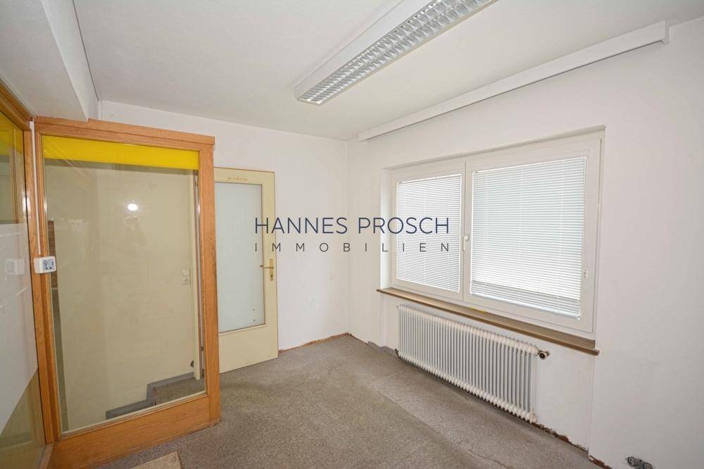 Kleiner Büroraum mit Fenster, Heizkörper und Zugang zu einem Treppenhaus, ausgestattet mit Teppichboden.