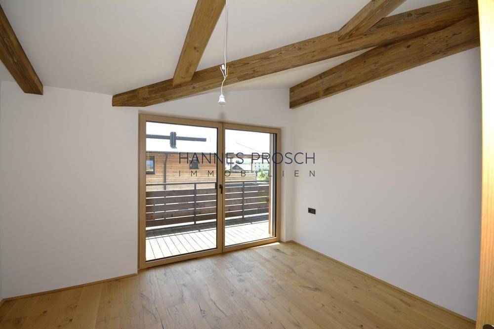 Lichtdurchflutetes Zimmer mit Holzboden, Holzbalken und Schiebetüren, die auf einen Balkon führen.