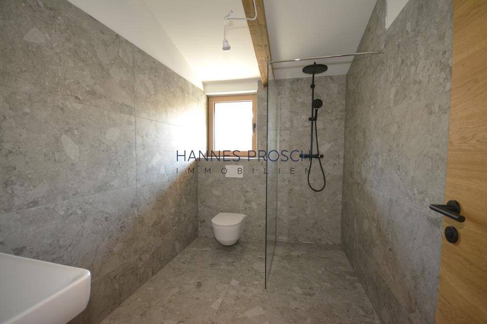 Modernes Badezimmer mit grauen Fliesen, wandhängendem WC und begehbarer Dusche mit Regenduschkopf.