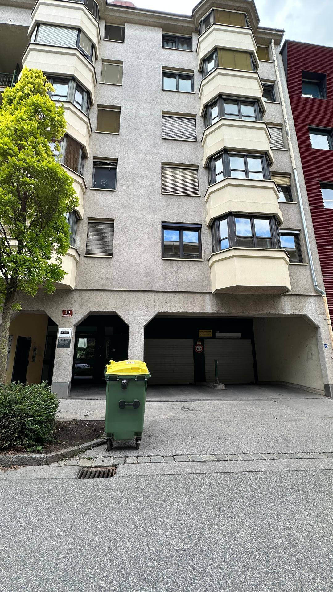 Fassade eines Mehrfamilienhauses mit integriertem Tiefgarageneingang und einem grünen Mülleimer.