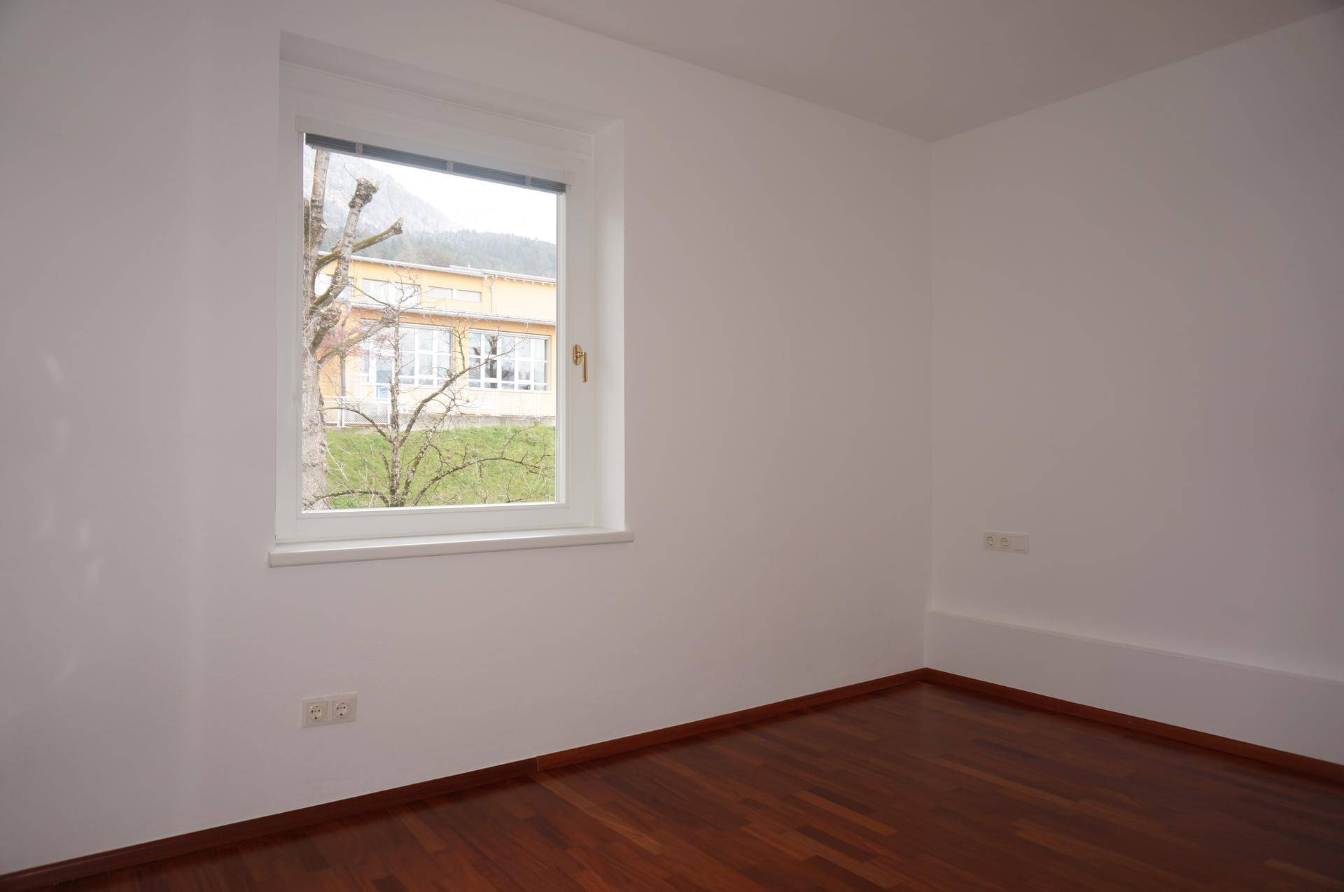 Helles, leeres Zimmer mit Holzboden und Fensterblick ins Grüne.