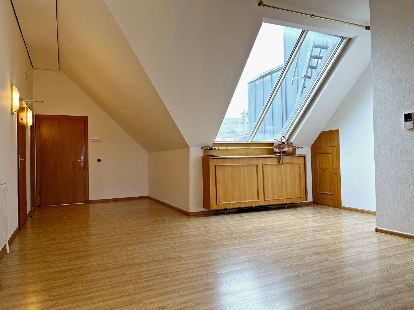 Leerer Raum mit Laminatboden, Holztür und großem schrägen Dachfenster mit Blick auf ein Gebäude.