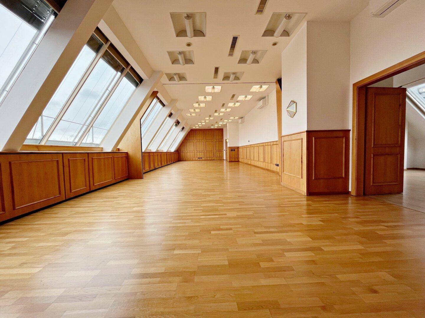 Großer, leerer Büroraum mit schrägen Fenstern, Holzvertäfelung und hellem Parkettboden.
