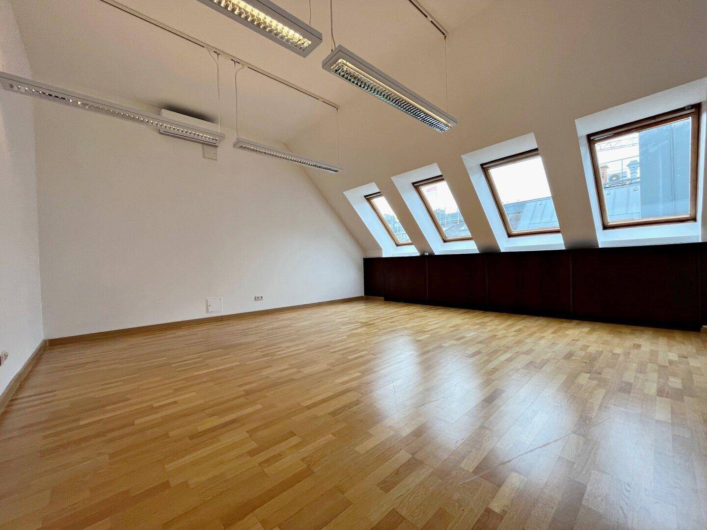 Leerer Raum mit Dachschrägen, drei Dachfenstern, dunkler Holzvertäfelung und hellem Holzboden.