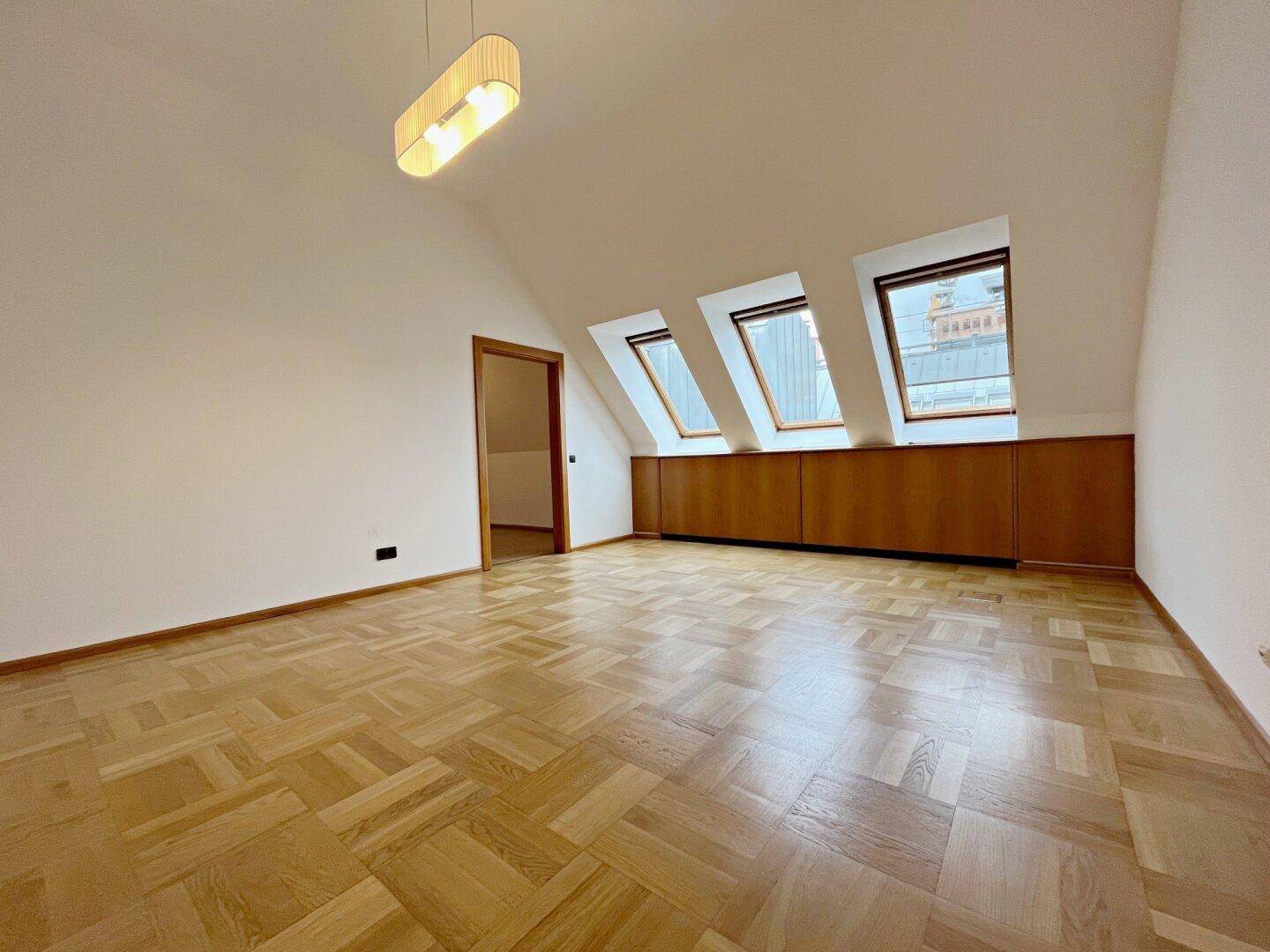 Leerer Raum mit Parkettboden, Dachschrägen, drei Dachfenstern und moderner Pendelleuchte.