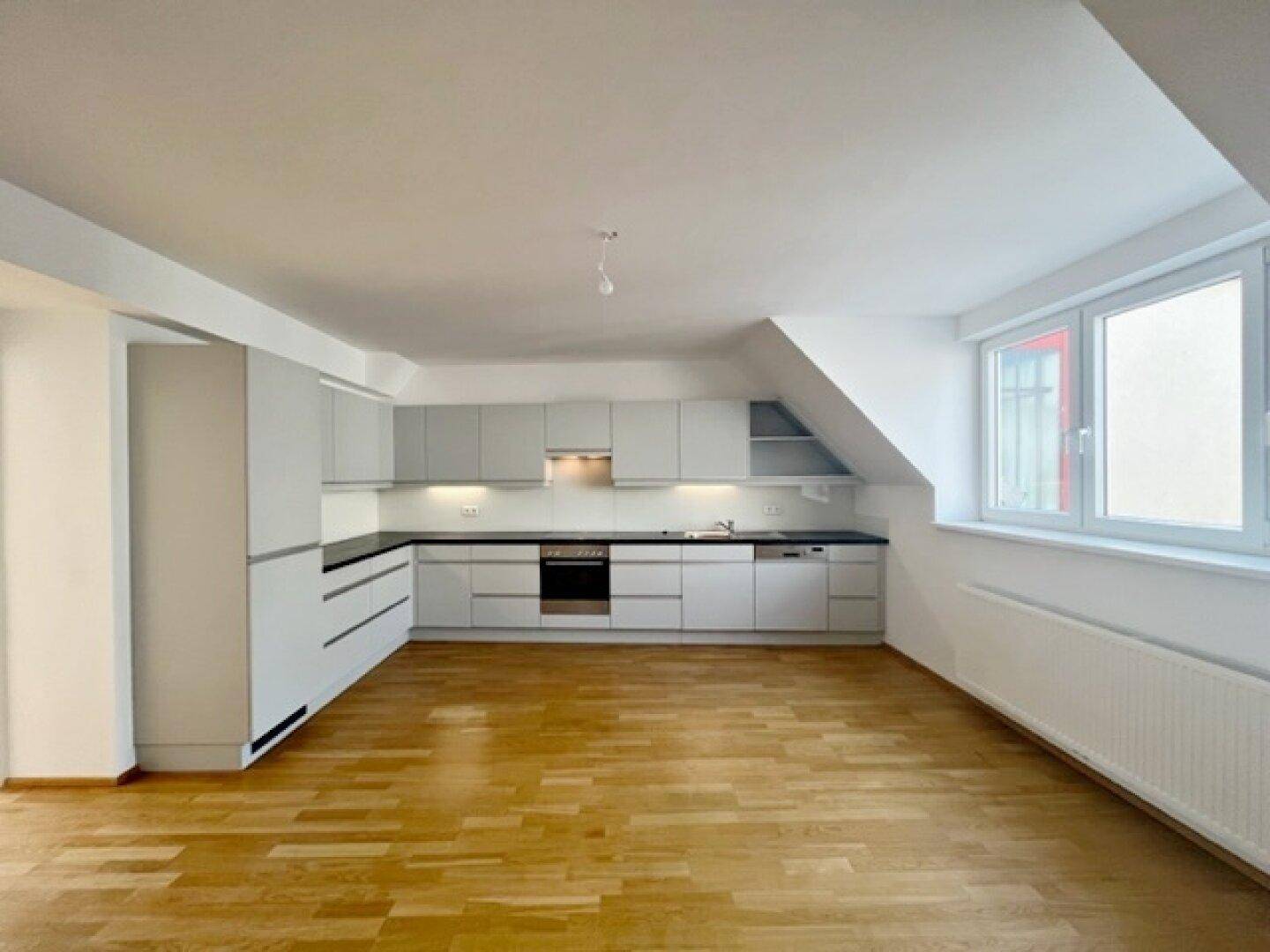 Helle Küche mit modernen grauen Schränken, dunkler Arbeitsplatte und Holzparkettboden unter einem Fenster.