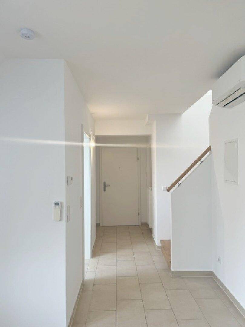 Heller Flur mit Fliesenboden, weiß gestrichenen Wänden und einer Klimaanlage an der Decke.