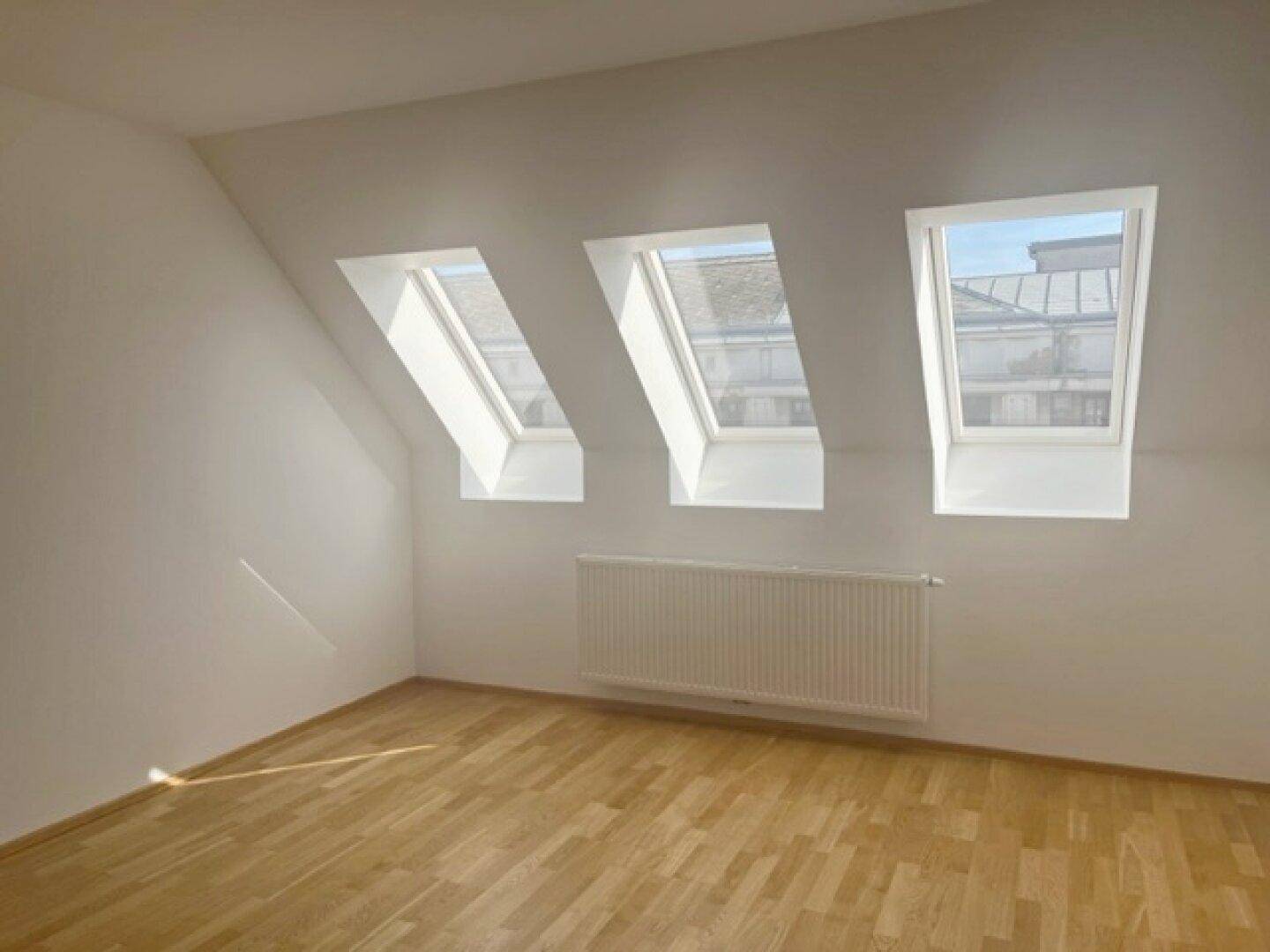 Dachgeschosszimmer mit schrägen Wänden, Holzparkett und drei großen Dachfenstern für natürliche Belichtung.