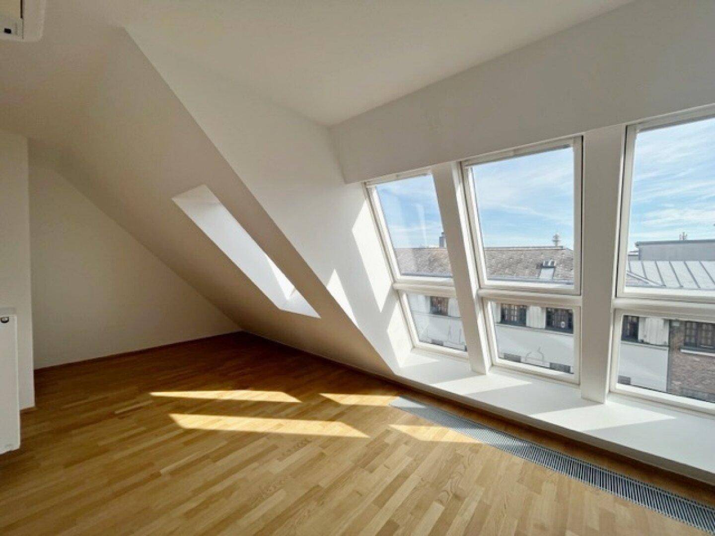 Heller Dachgeschossraum mit schrägen Wänden, Holzparkett und einer breiten Fensterfront mit Stadtblick.