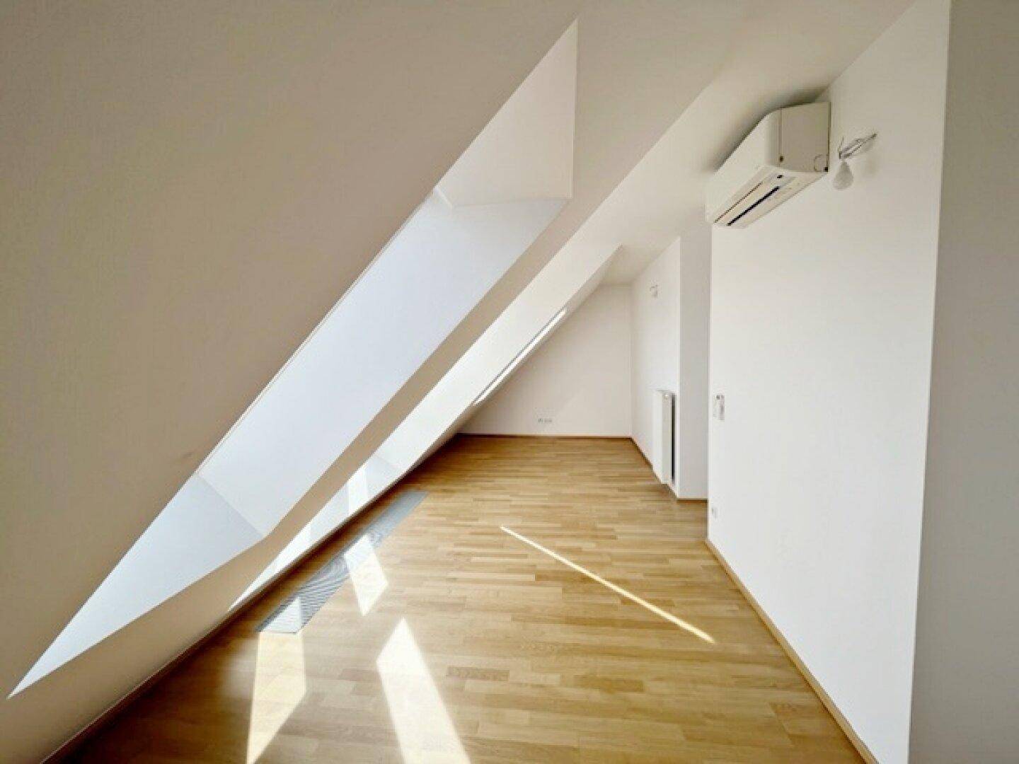Langer, heller Raum mit Holzparkettboden, schrägen Wänden und einer Klimaanlage an der Decke.