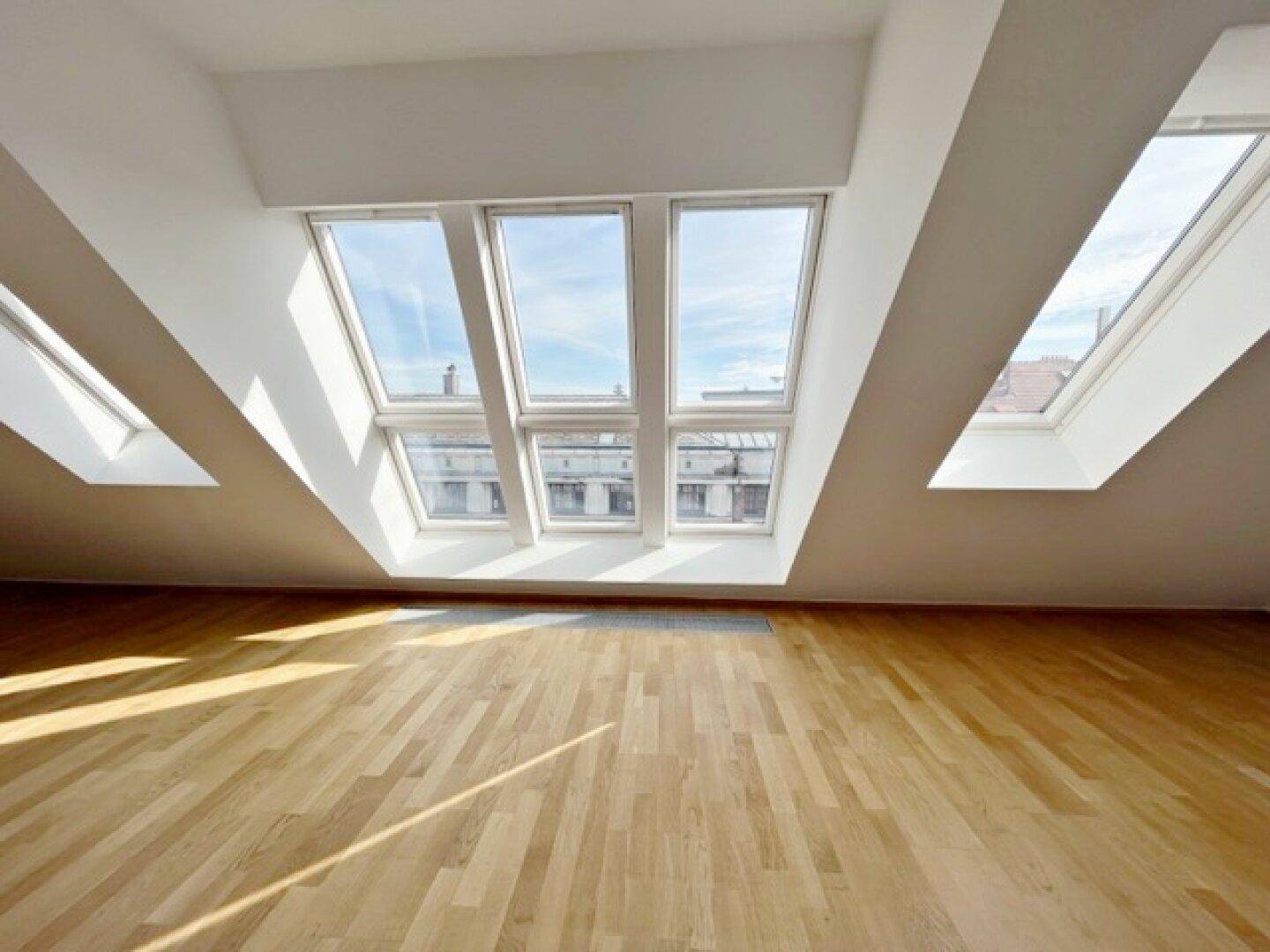 Geräumiger Dachgeschossraum mit breiter Fensterfront und Holzparkettboden, der viel Sonnenlicht empfängt.