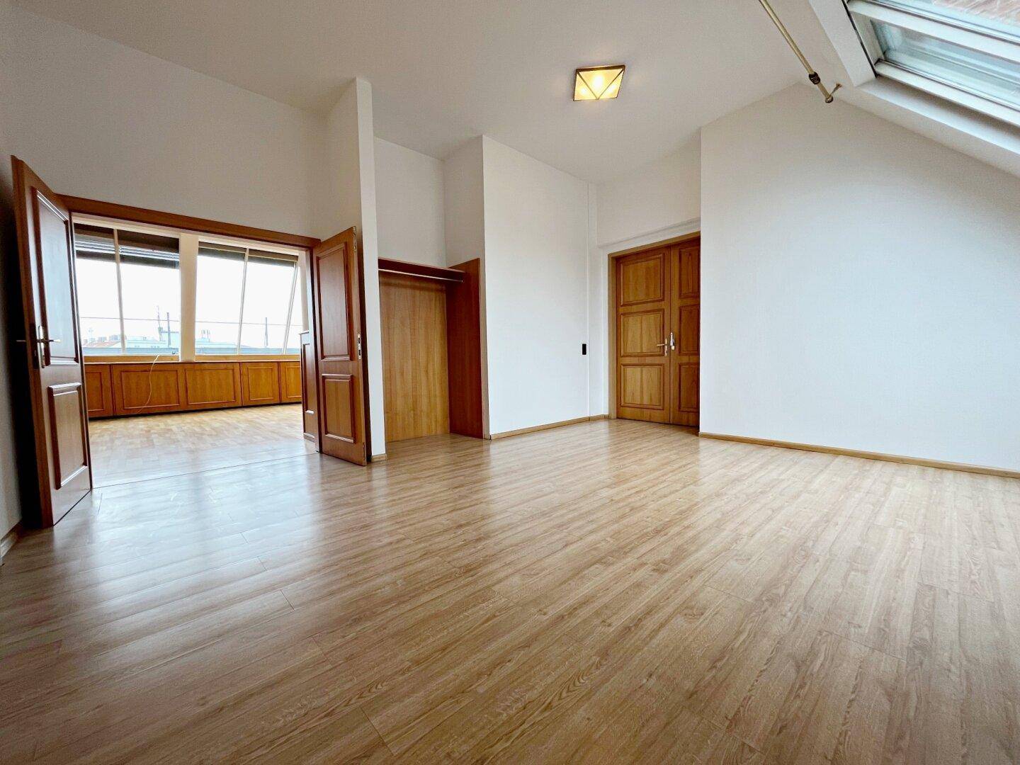 Leerer Raum mit Laminatboden, weißen Wänden, Holztüren und einem Dachfenster, hell und geräumig.