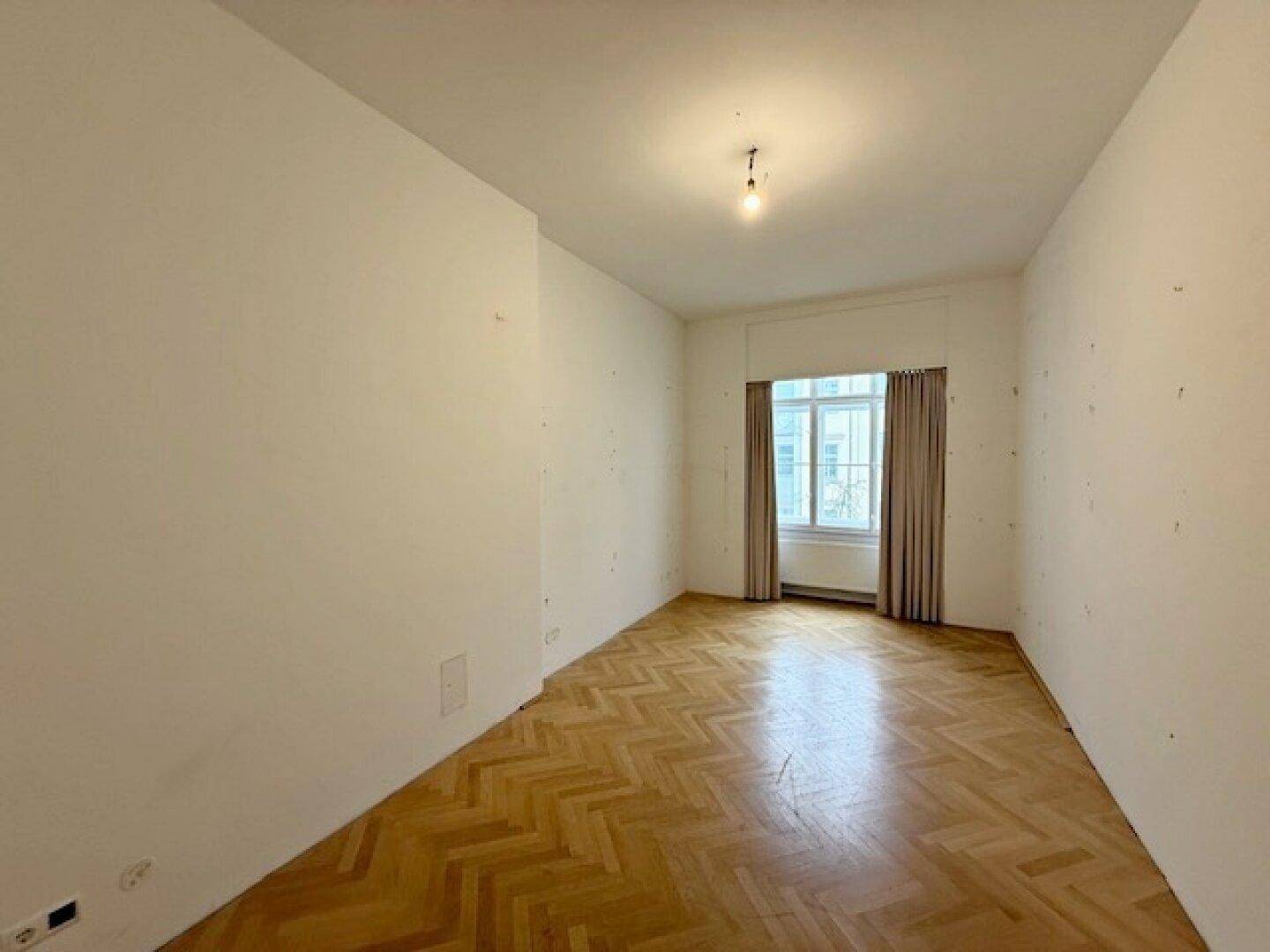 Unmöblierter Raum mit Fischgrätparkett und Fenster mit Vorhängen, bereit zur Nutzung.