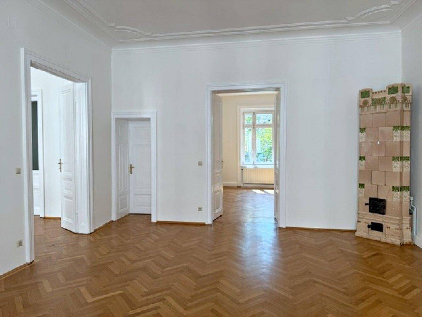 Geräumiges Wohnzimmer mit Fischgrätparkett und einem traditionellen Kachelofen als Blickfang.