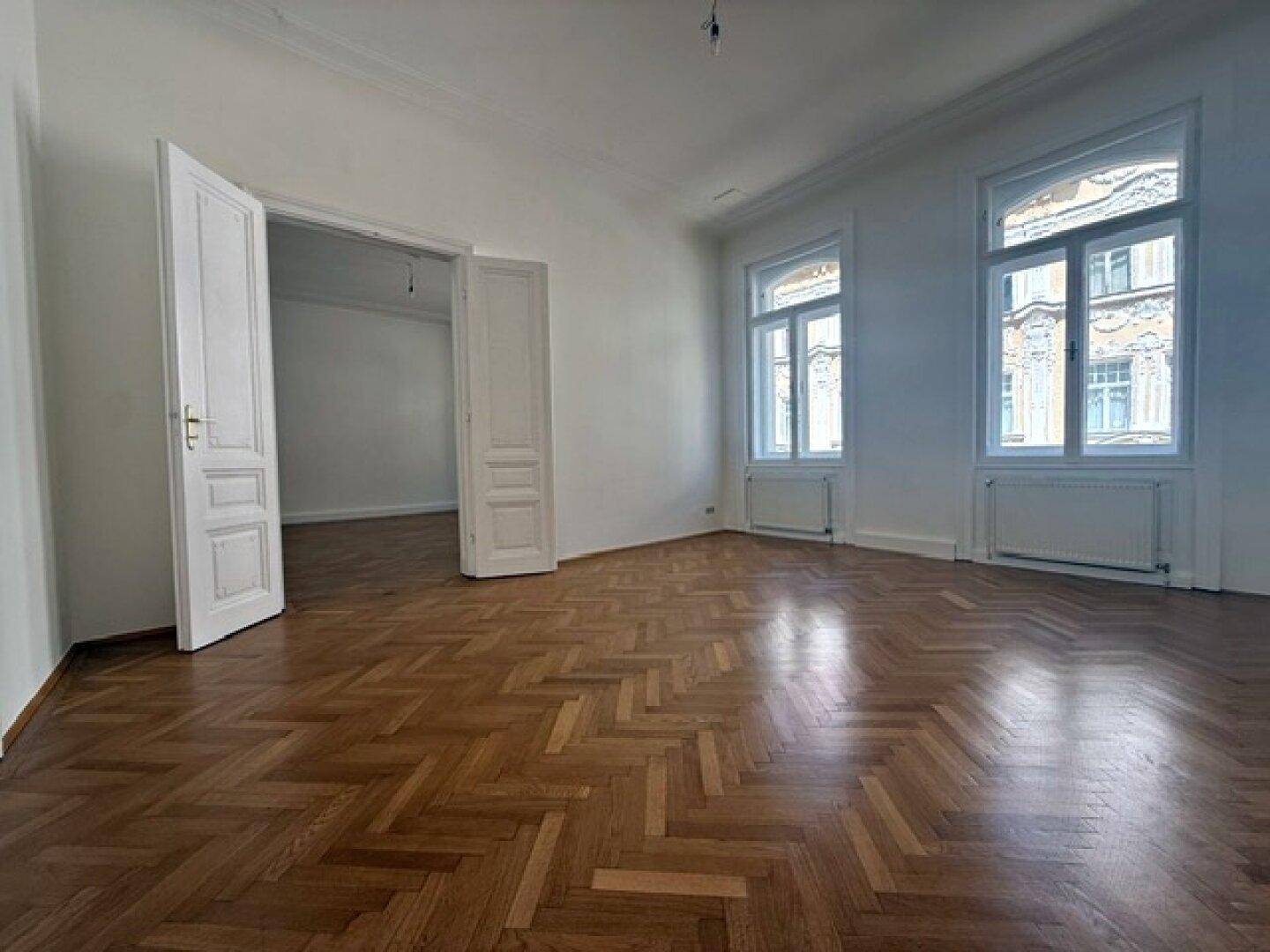 Helles Wohnzimmer mit Fischgrätparkett, hohen Decken und großen Fenstern, die viel Tageslicht hereinlassen.