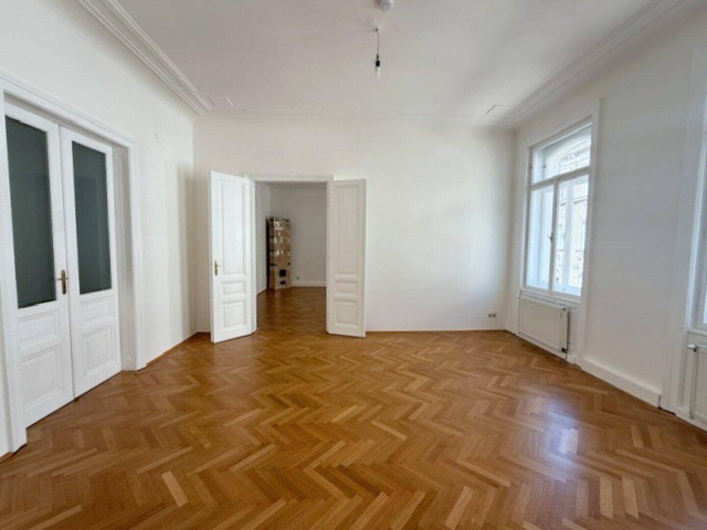 Helles Wohnzimmer mit Fischgrätparkett und hohen Doppeltüren, die zu weiteren Räumen führen.