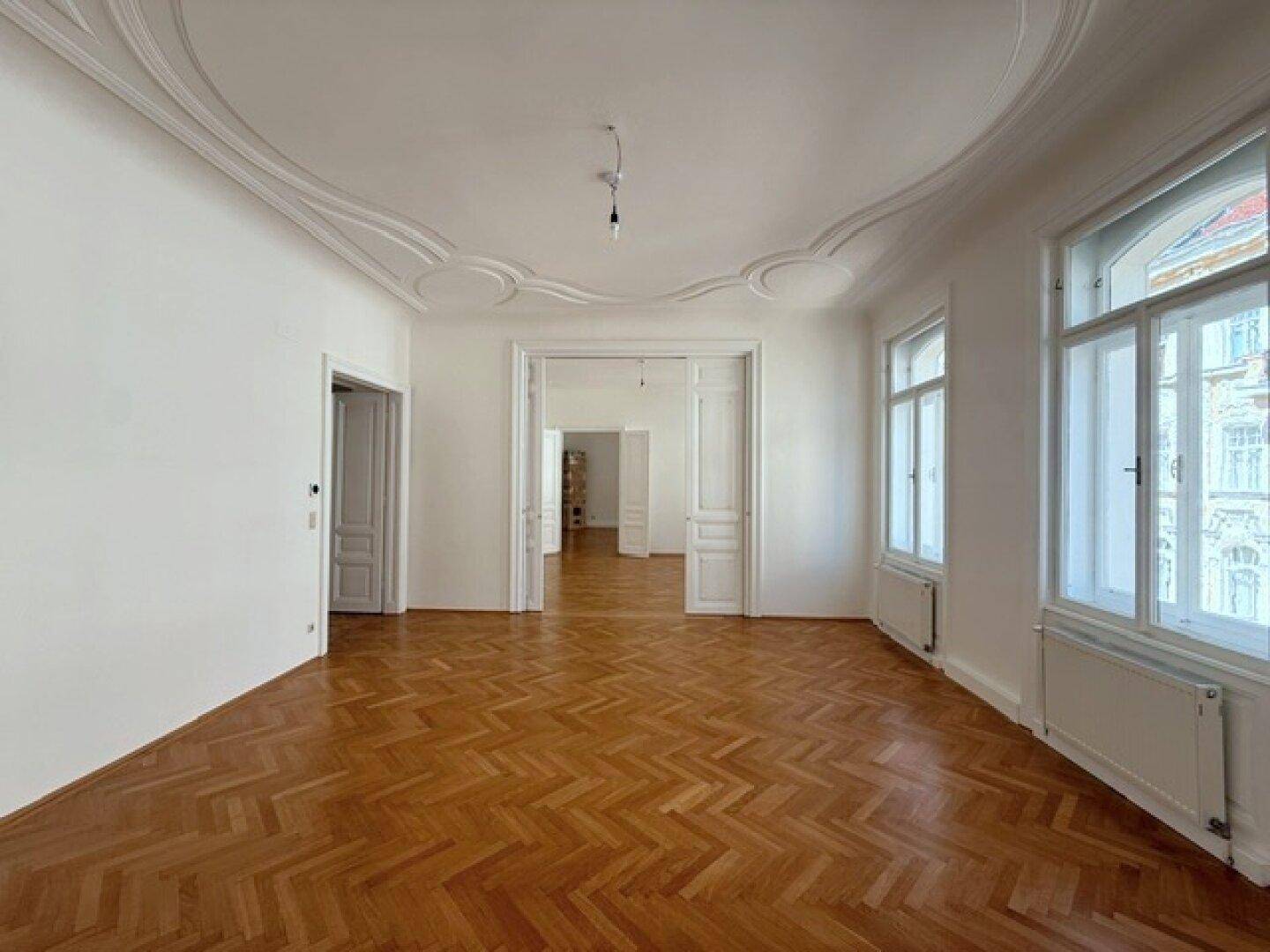 Weitläufiges Wohnzimmer mit Fischgrätparkett, Stuckdecken und hohen Fenstern, die eine helle Atmosphäre schaffen.