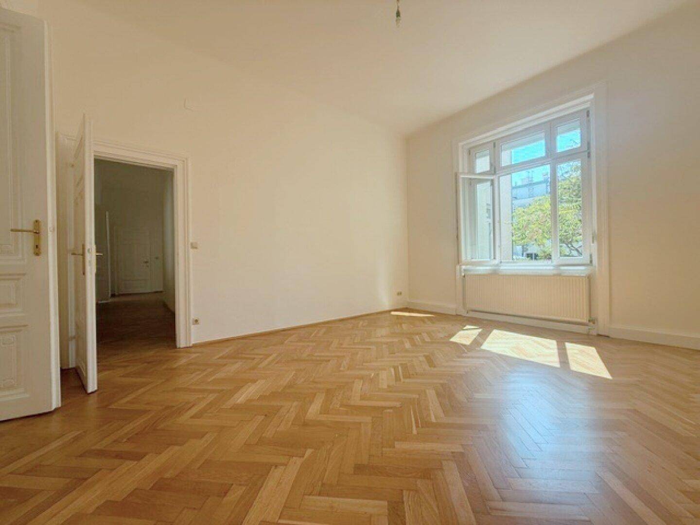 Helles Zimmer mit Fischgrätparkett und einem großen Fenster, das viel Sonnenlicht in den Raum bringt.