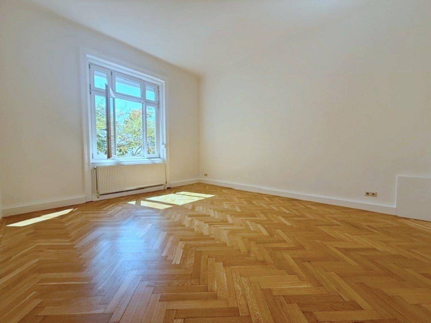 Sonniger Raum mit Fischgrätparkett und einem großen Fenster, das den Blick nach draußen freigibt.