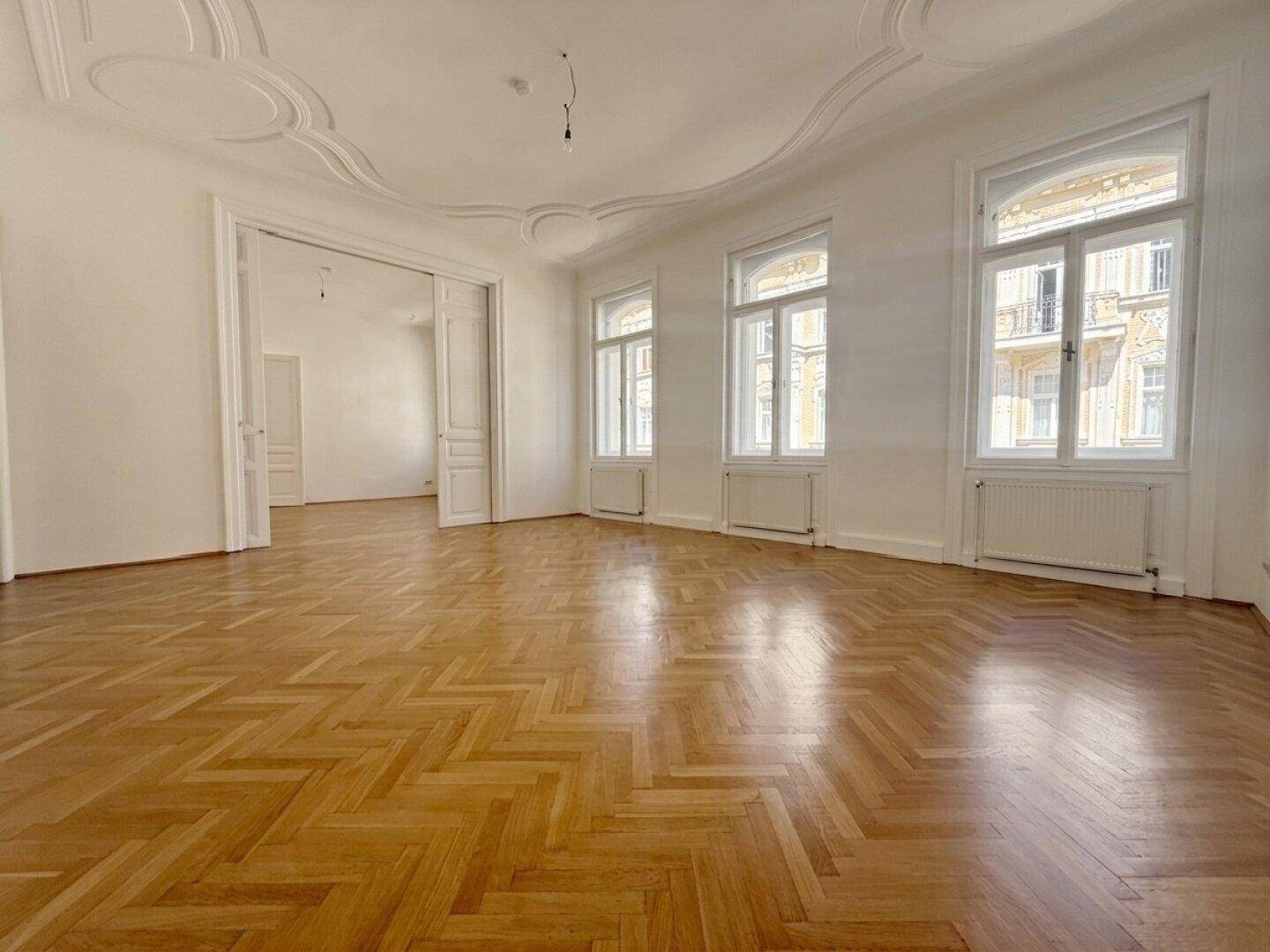 Helles Altbau-Wohnzimmer mit elegantem Fischgrätparkett, großen Fenstern und dekorativem Deckenstuck.