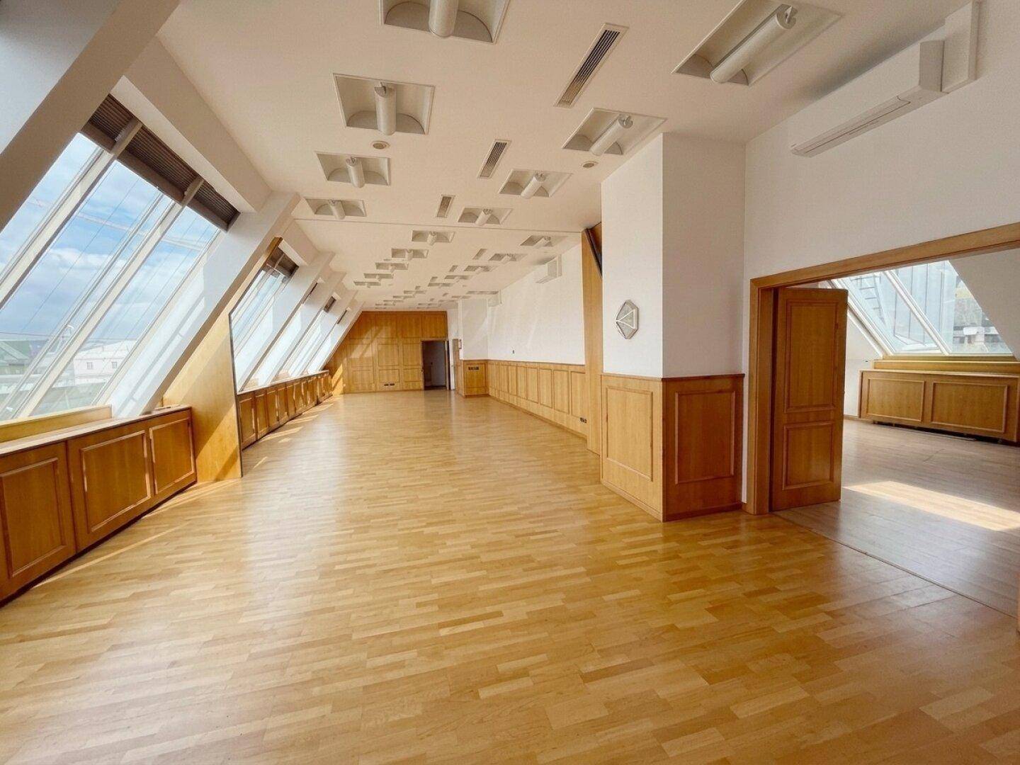 Geräumiger, leerer Büroraum mit schrägen Fenstern, Holzvertäfelung und hellem Holzparkettboden.