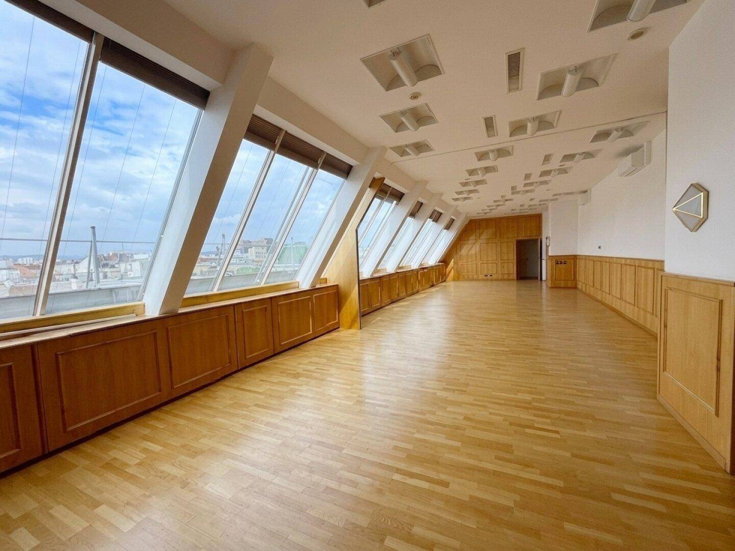 Heller, leerer Büroraum mit schrägen Fenstern, Holzvertäfelung und Blick auf die Stadt.