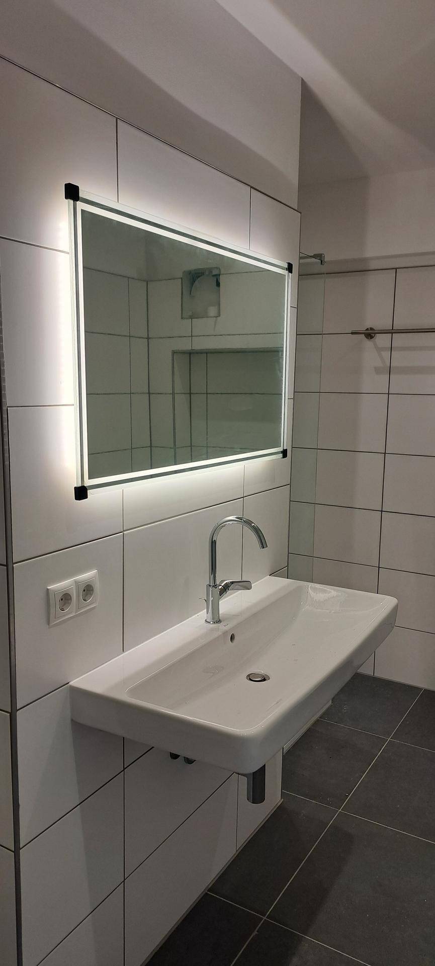 Modernes Badezimmer mit beleuchtetem Spiegel, Waschtisch und großformatigen weißen Wandfliesen.