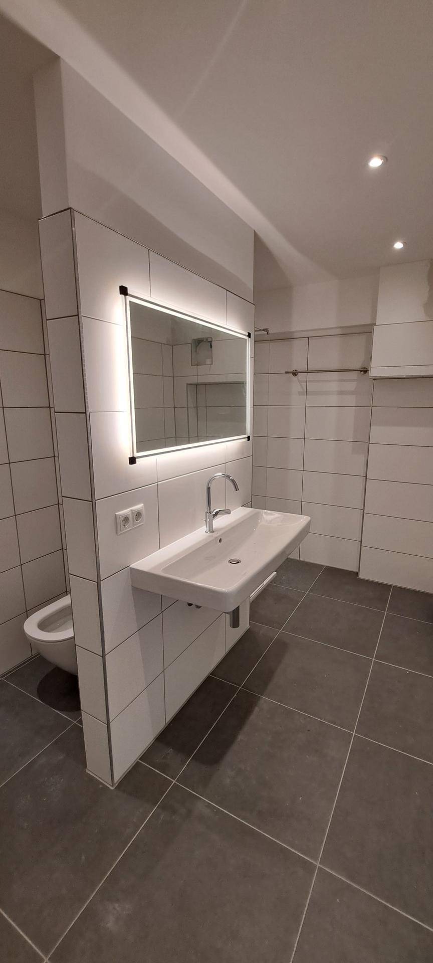 Zeitgemäßes Badezimmer mit Waschtisch, beleuchtetem Spiegel und wandhängendem WC auf Fliesenboden.