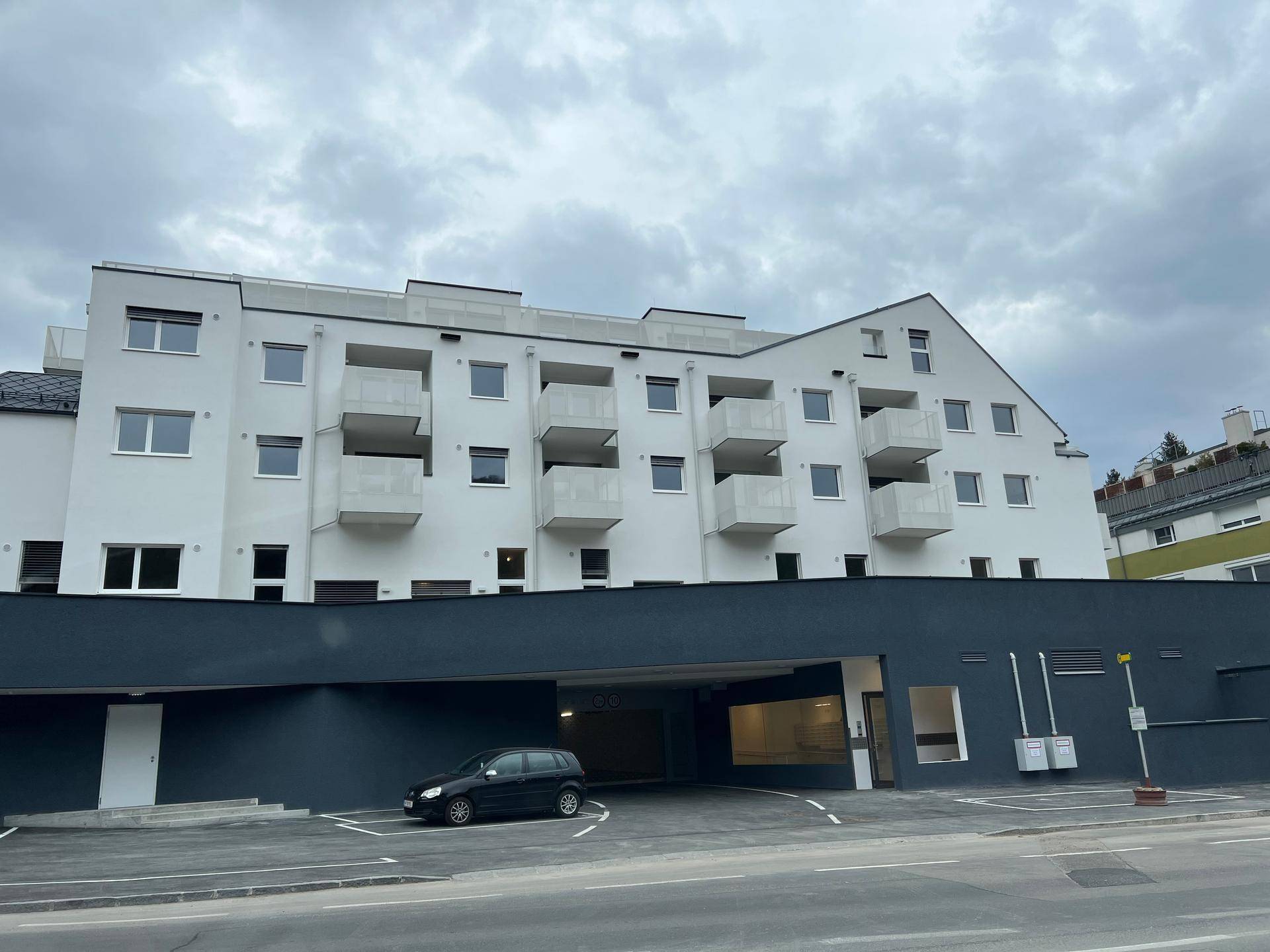 Moderne Wohnanlage mit weißer Fassade, Balkonen und Tiefgaragenzufahrt unter bewölktem Himmel.