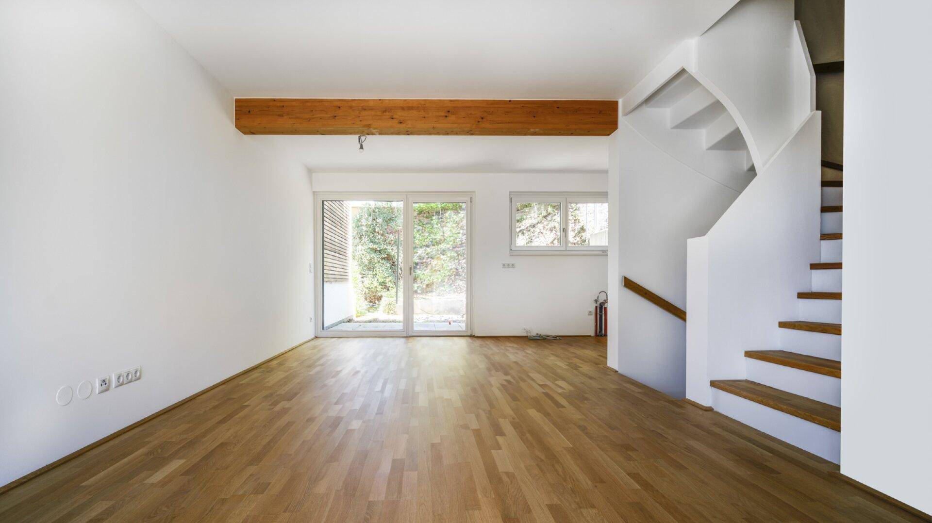 Helles, leeres Wohnzimmer mit Holzbalken, Parkettboden und Zugang zum Garten.
