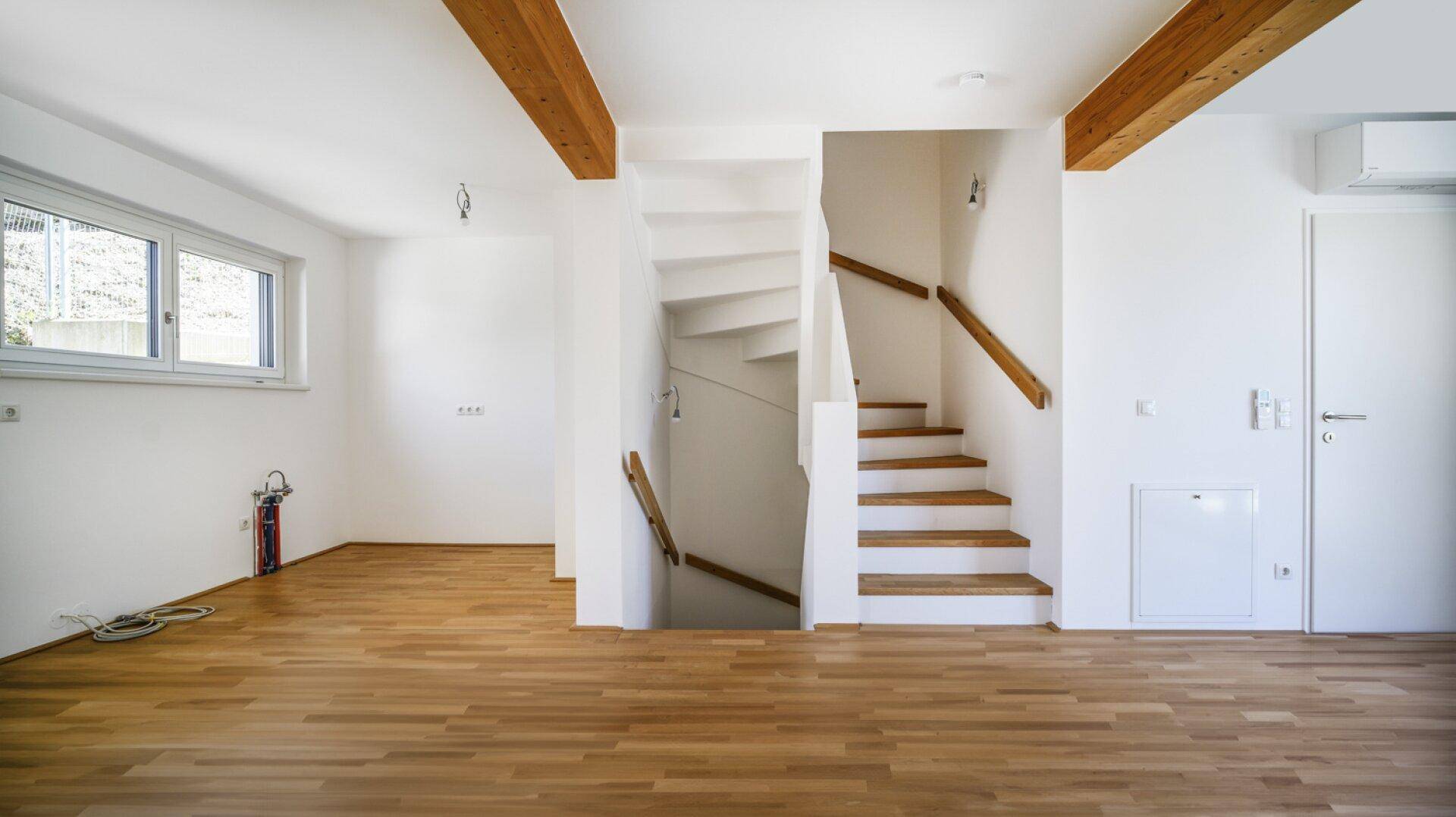 Heller Wohnbereich mit Parkettboden, sichtbaren Holzbalken und Treppe in die obere Etage.