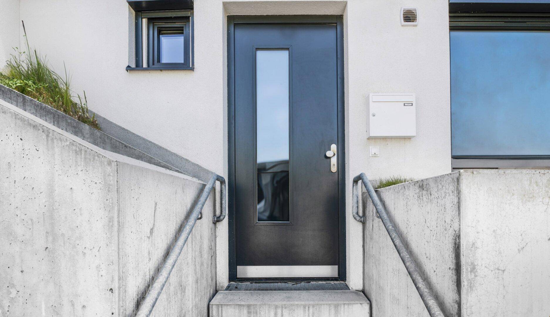 Moderne schwarze Eingangstür mit Glasausschnitt und Edelstahlgriffen, umgeben von Betonstufen.