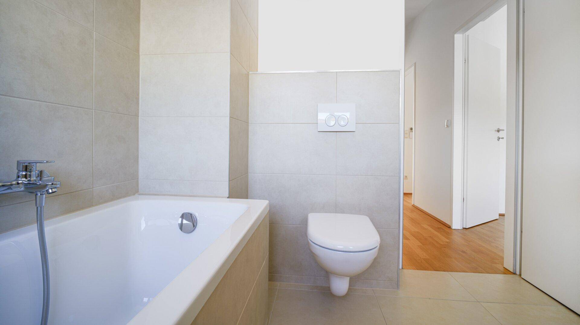 Helles Badezimmer mit Badewanne, wandhängendem WC und beigefarbenen Wand- und Bodenfliesen.