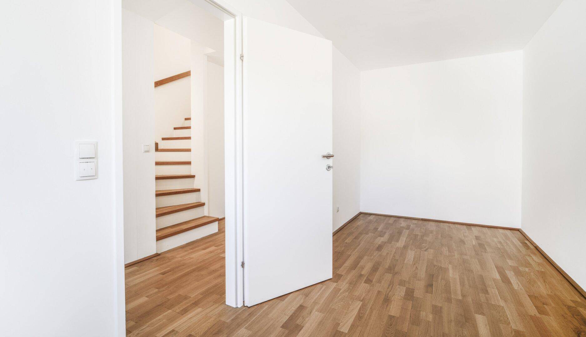 Leeres Zimmer mit Parkettboden und Blick auf die Holztreppe durch die offene Tür.