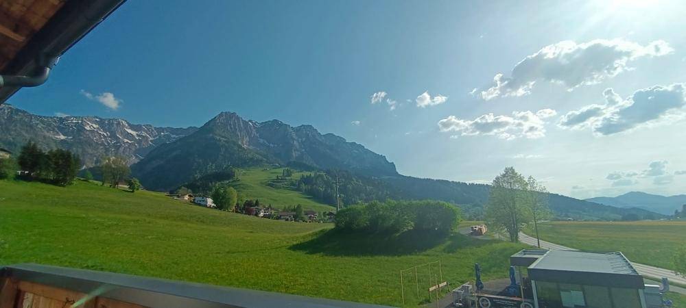 Sonniger Ausblick von einem Balkon auf grüne Wiesen und die umliegende Bergwelt.