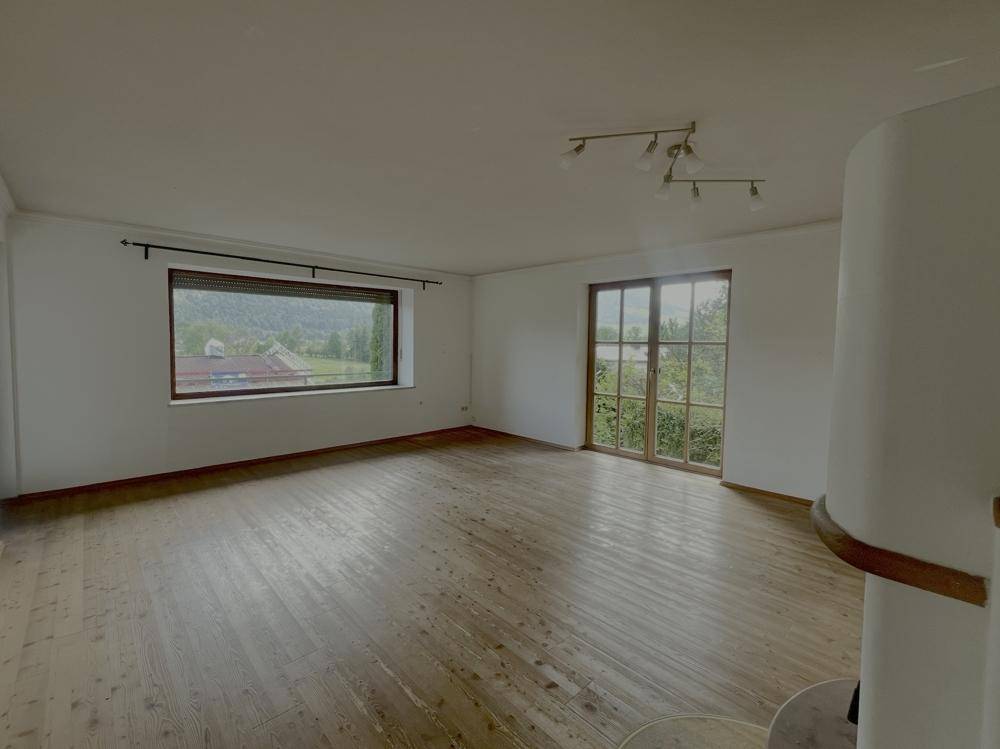 Lichtdurchflutetes Wohnzimmer mit Holzboden und zwei Fenstern, die einen Blick ins Grüne ermöglichen.