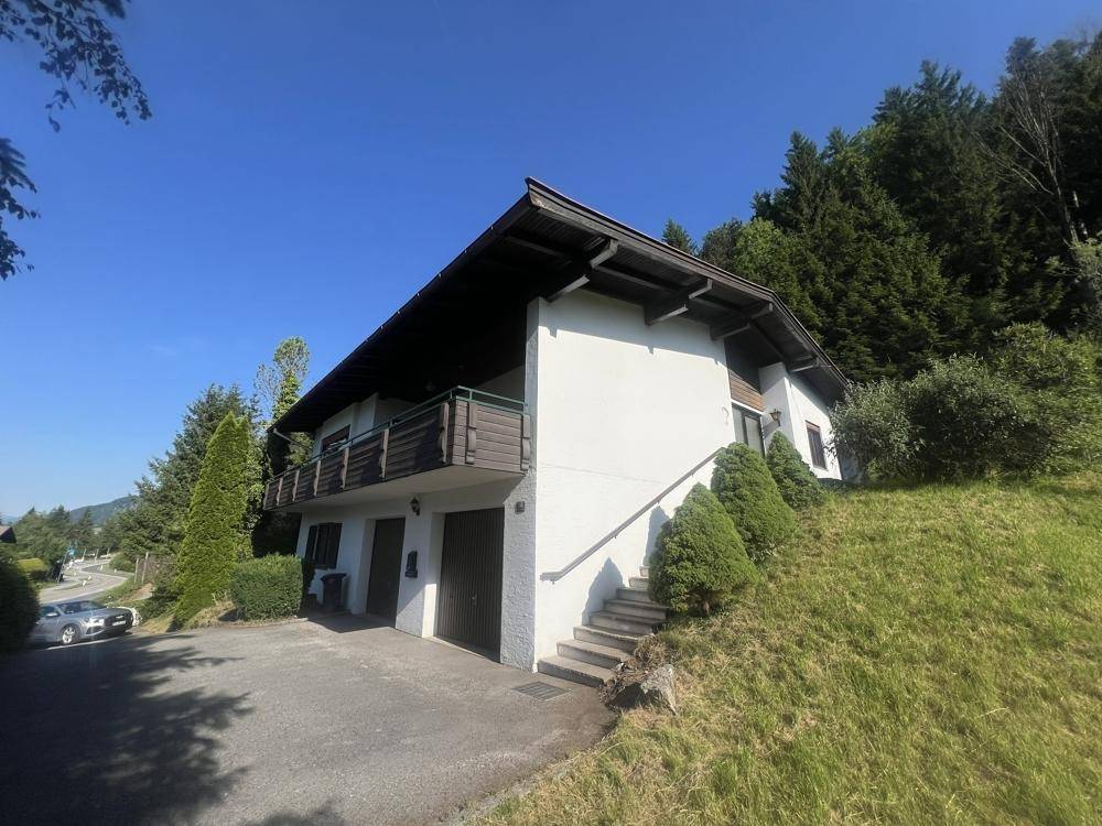 Hausfassade mit Garagen und einer Treppe, eingebettet in eine grüne Hanglage unter blauem Himmel.