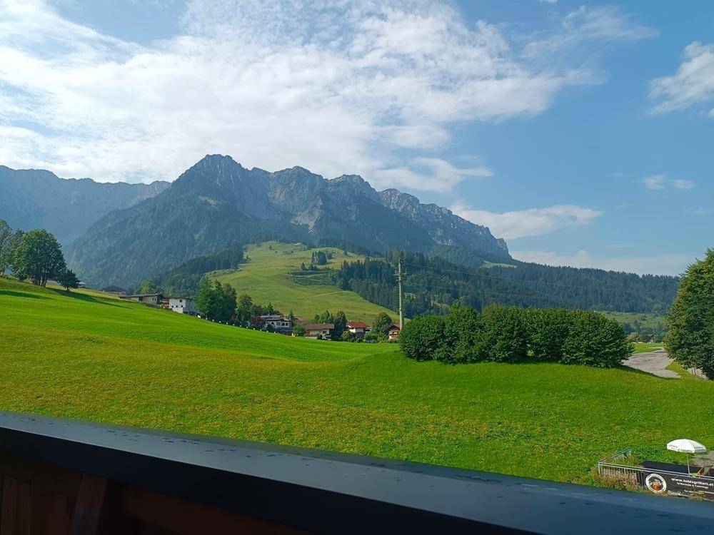 Panoramablick von einem Balkon auf grüne Wiesen und majestätische Berge unter blauem Himmel.