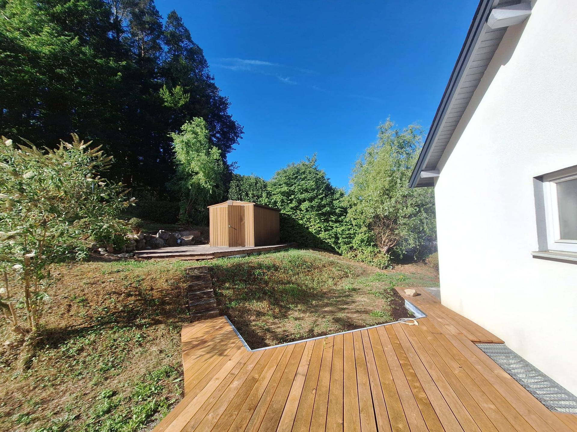 Sonniger Gartenbereich mit einer Holzterrasse und einem kleinen Geräteschuppen am Waldrand.