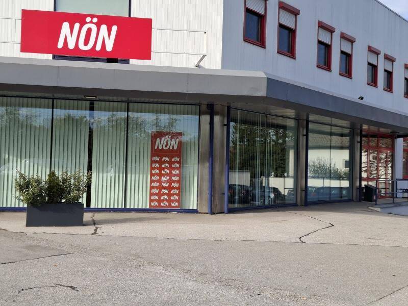 Außenansicht eines modernen Geschäftsgebäudes mit großen Schaufenstern, rotem Logo und einer Zufahrt.